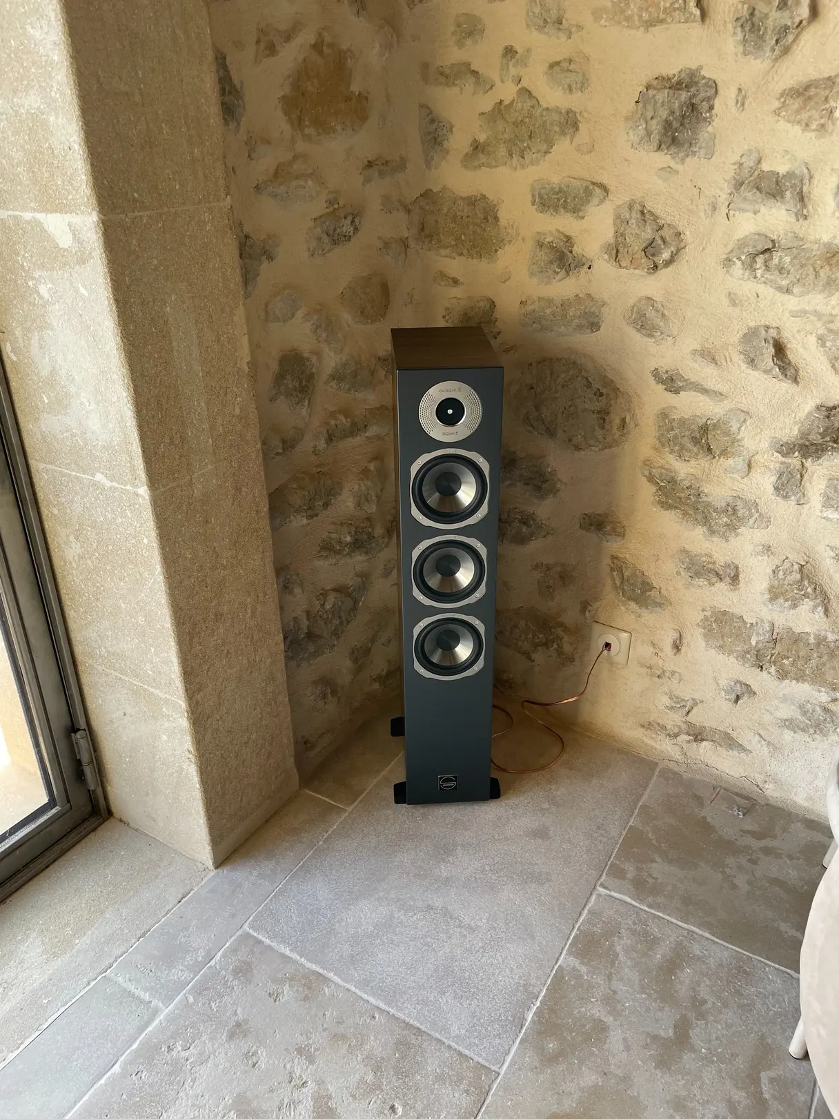 Enceinte colonne installée dans un angle, contre un mur en pierre apparente