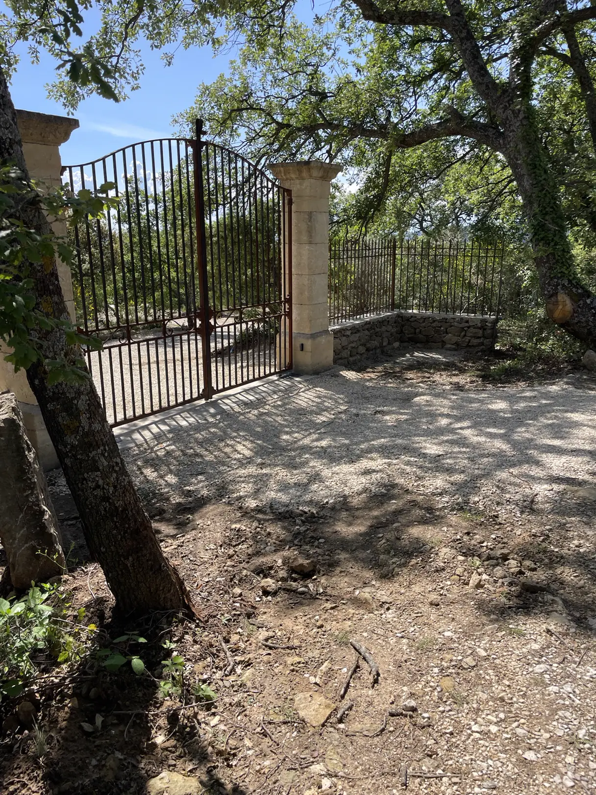 Portail en fer forgé entre deux piliers de pierre, sous les chênes, à l'entrée d'un domaine provençal
