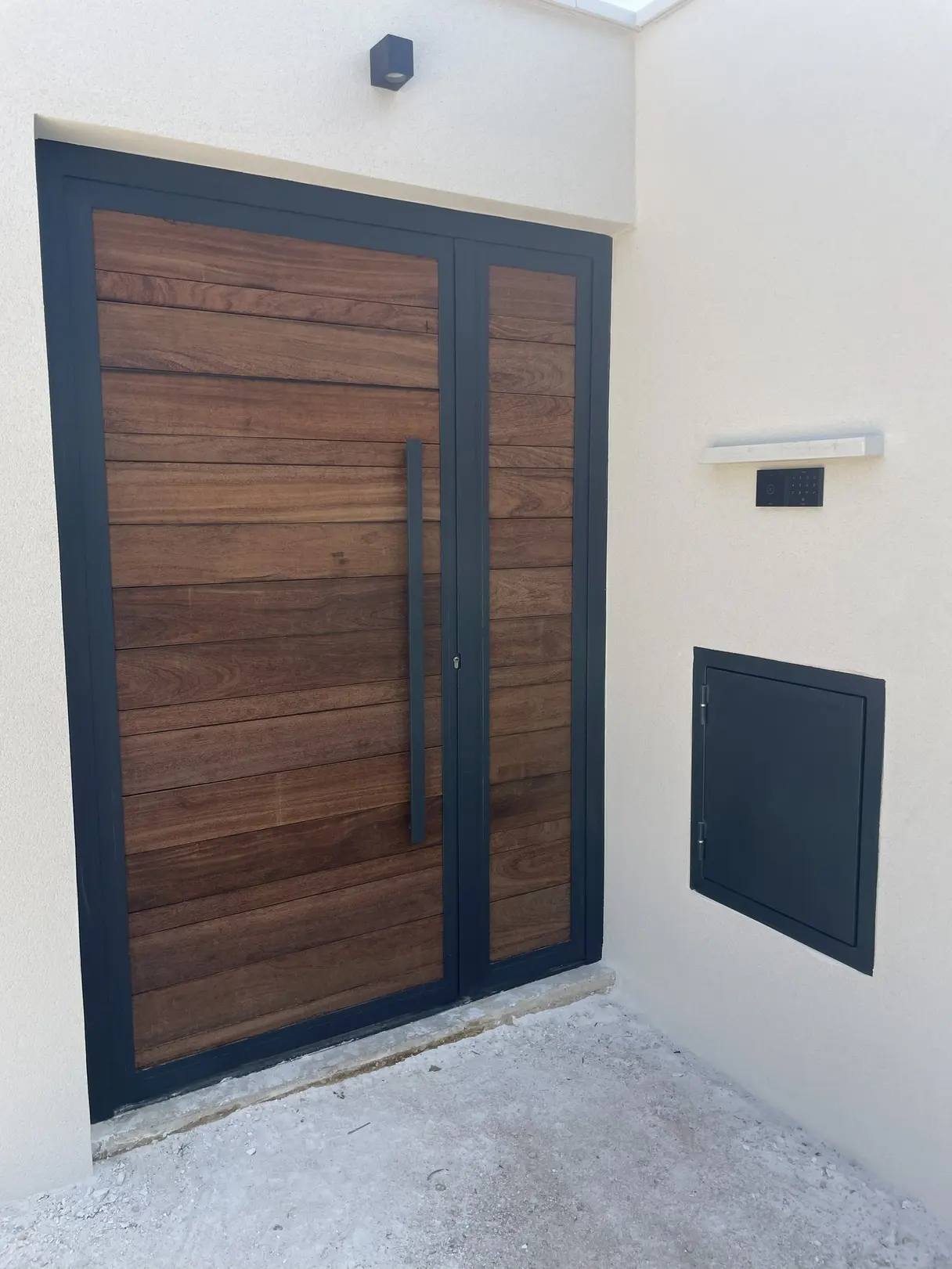 Porte d'entrée en bois et acier avec platine de contrôle d'accès et trappe à colis intégrée dans le mur